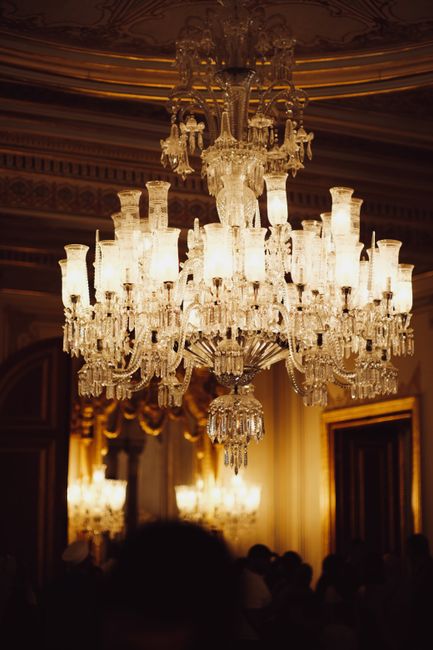 A large, ornate crystal chandelier hangs from the ceiling of an opulent room, its multiple tiers of glass prisms and candle-style lights illuminated. The chandelier is centrally positioned, with detailed carvings on the ceiling's decorative molding visible in the background. Below the chandelier, blurred figures of people can be seen, suggesting a formal event or gathering. The room's elegant decor includes gold accents, draped curtains, and tall mirrors. During house removals or relocation processes, similar chandeliers may need careful handling when moving furniture, and the scene underscores the importance of professional packing and transport services provided by companies like Removals Crystal Palace to ensure delicate fixtures are safely preserved during home relocation or moving logistics.