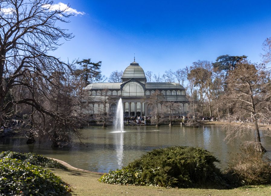 A large glasshouse or conservatory with a domed roof and arched windows is situated behind a pond in a park setting. The pond has a central fountain with water spraying upward, and the area around it is landscaped with low green bushes and bare trees, indicating a cold season. There are pedestrians visible near the glasshouse, and the sky above is clear with a few clouds. The scene is illuminated by natural daylight, showing the building's structure and the surrounding environment clearly. The photograph captures the exterior view in a peaceful park used for leisure and outdoor activities, and it may be part of a house removal or relocation project involving the transportation of furniture and equipment to or from this property, as managed by Removals Crystal Palace.