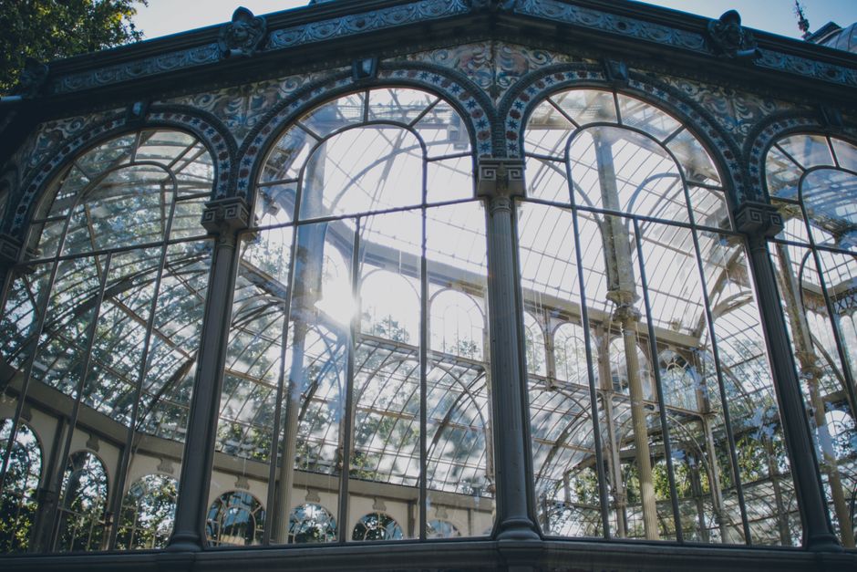 A detailed view of an ornate glass and metal greenhouse structure with large, arched windows and decorative columns, allowing sunlight to filter through and illuminate the interior. The greenhouse features curved glass panels supported by metal framing, with visible reflections on the glass surfaces. Trees and foliage are partially visible through the transparent windows, suggesting an outdoor garden setting. This type of environment is commonly used in conservatories or botanical displays, often associated with gardens like those in Crystal Palace. In the context of house removals and furniture transport, such a structure could be an example of delicate glasswork requiring careful handling during a home relocation process, supported by companies like Removals Crystal Palace specializing in packing and moving fragile fixtures or garden features.
