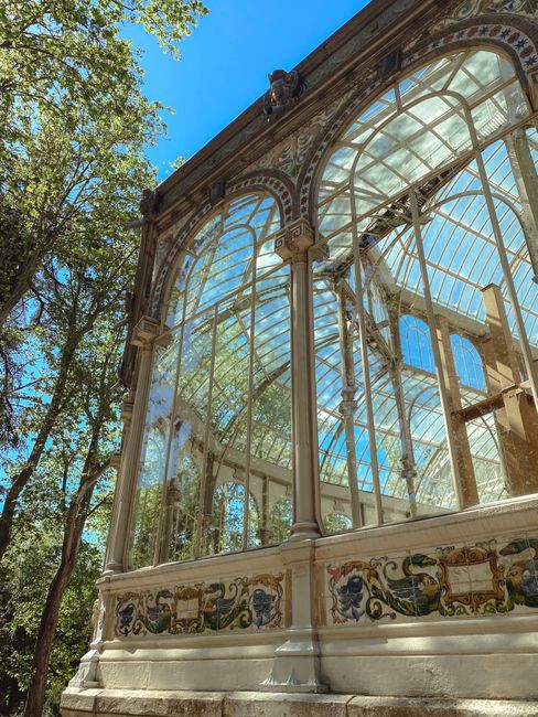 A large glasshouse or conservatory with a domed roof and arched windows is situated behind a pond in a park setting. The pond has a central fountain with water spraying upward, and the area around it is landscaped with low green bushes and bare trees, indicating a cold season. There are pedestrians visible near the glasshouse, and the sky above is clear with a few clouds. The scene is illuminated by natural daylight, showing the building's structure and the surrounding environment clearly. The photograph captures the exterior view in a peaceful park used for leisure and outdoor activities, and it may be part of a house removal or relocation project involving the transportation of furniture and equipment to or from this property, as managed by Removals Crystal Palace.