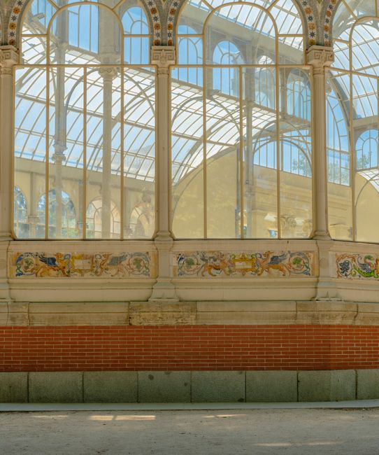 Photograph of the interior of a historic conservatory or greenhouse with large, arched glass windows supported by ornate metal framing painted in a light color. The upper section features multiple panes of clear glass allowing natural light to fill the space, with visible metal decorative elements and a curved roof structure. Below the windows, there is a decorative frieze or mural depicting stylized, colorful figures and patterns. The lower part of the image shows a brick wall base and a concrete or stone ledge running along the bottom. The scene reflects an elegant architectural design typical of grand Victorian or Edwardian greenhouses, with natural light illuminating the interior, which could be part of a home or public garden. This setting may relate to the process of home relocation or furniture transport during a modern house move, as managed by the professional team at Removals Crystal Palace.